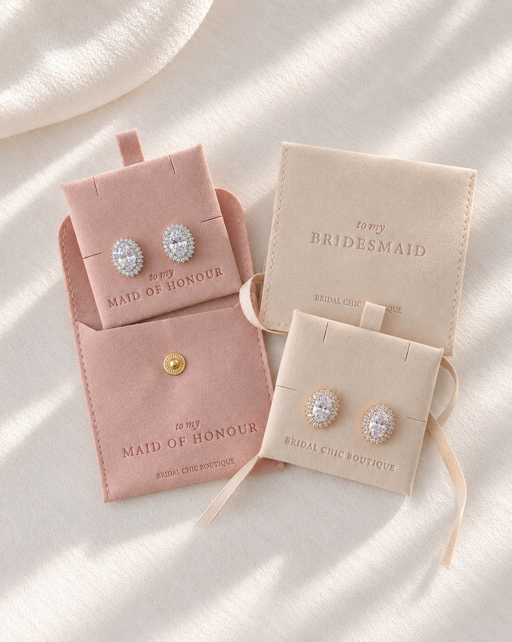 Three velvet jewelry display cards in blush pink and cream beige, each holding a pair of oval stud earrings with clear stones and a gold-tone setting, arranged on a softly lit white fabric background. The cards are printed with bridal/bridesmaid event text.