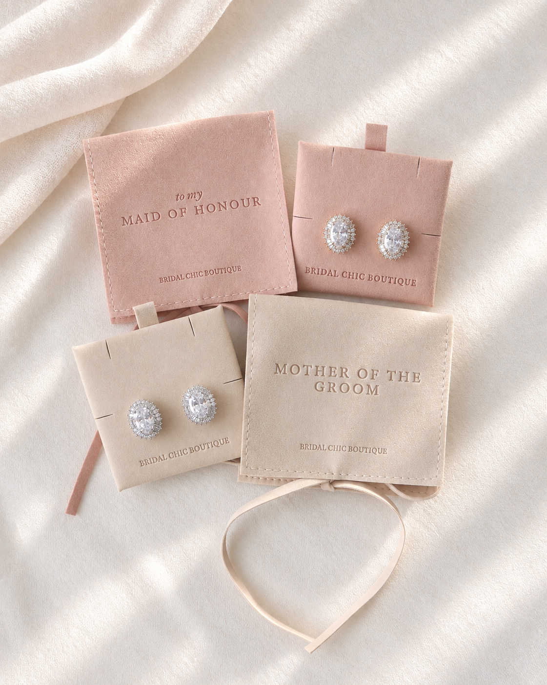 Three pairs of silver-toned stud earrings displayed on blush and cream jewelry cards, arranged on a soft white fabric background with a cream headband in front.