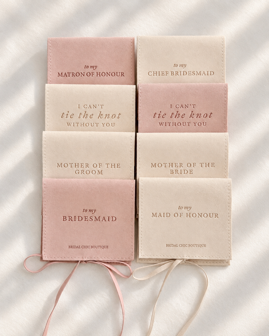 Six blush pink and cream fabric wedding favor envelopes with embroidered text, arranged in a grid on a white textured fabric background with delicate ribbon ties.