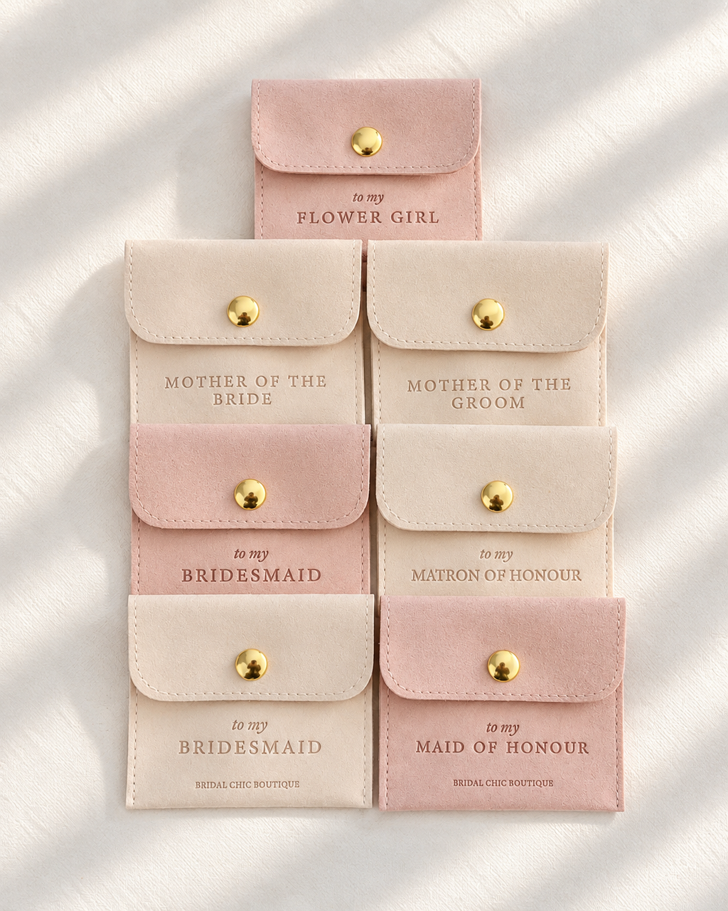 Six small fabric pouches with gold snap closures are arranged on a white textured surface with soft shadows. The pouches come in blush pink and cream tones and are printed with different wedding party titles including Flower Girl, Mother of the Bride, Bridesmaid, Matron of Honour, and Maid of Honour.