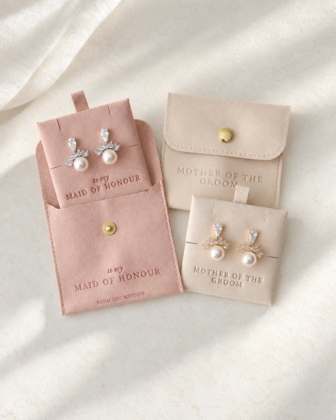 Two jewelry display pouches in blush pink and light beige, each holding matching pearl stud earrings with clear crystal floral tops, arranged on a soft white fabric background.